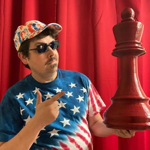 Op de afbeelding: Een persoon met een rood, wit en blauwe tie-dye baseballpet en zonnebril houdt een groot, rood, houten schaakstuk vast. Het schaakstuk is een dame.