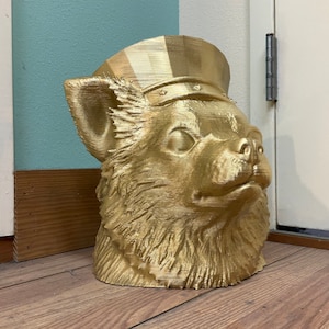 May include: A gold-painted 3D printed bust of a chihuahua wearing a gold-painted hat. The bust is sitting on a wooden floor.