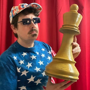 May include: A person wearing a red, white, and blue tie-dye shirt and a red, white, and blue baseball cap is holding a large, yellow wooden chess piece. The chess piece is a queen.