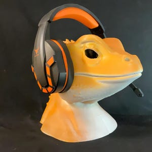 Op de afbeelding: Een feloranje en zwarte gaming-headset met een microfoon wordt gedragen door een geel en wit keramiek beeldje van een hagedis.