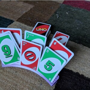 May include: A purple card holder with a stack of UNO cards, including a green card with the number 9, a red card with the word "Skip", a green card with the number 5, and a red card with the number 4.