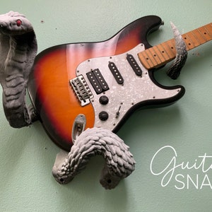 以下が含まれることがあります： ギターは、蛇の形をしたギタースタンドを使って壁に取り付けられています。スタンドは灰色のプラスチック製で、リアルな蛇のデザインになっています。ギターはサンバーストのエレキギターで、白いピックガードが付いています。壁のギターの下には、「guitar SNAKE」という文字が筆記体で書かれています。