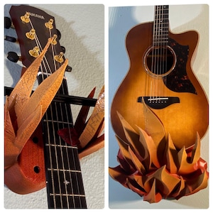 Puede incluir: Una guitarra acústica marrón con una golpeador negro y clavijas doradas está montada en una pared con un soporte en forma de llama hecho de material naranja y marrón. La guitarra tiene la palabra "YAMAHA" en la pala.