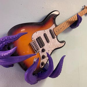 Peut inclure: Une guitare électrique avec une finition sunburst, un pickguard blanc et des micros noirs est ornée de tentacules violets. Les tentacules enroulent la guitare, créant une décoration murale unique. La guitare est fixée au mur.