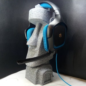 以下が含まれることがあります： 黒と青のゲーミングヘッドセット。イヤーカップに金のGロゴが付いています。ヘッドセットは灰色のスタンドの上にあります。