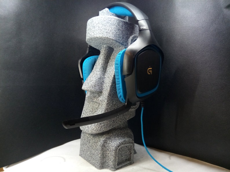 以下が含まれることがあります： 黒と青のゲーミングヘッドセット。イヤーカップに金のGロゴが付いています。ヘッドセットは灰色のスタンドの上にあります。