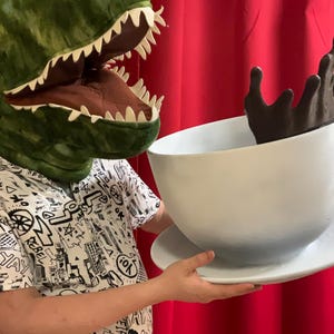 May include: A person wearing a green dinosaur head is holding a large white teacup with a brown liquid splashing out of it. The teacup is on a white saucer.