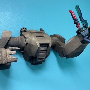 Può includere: Un supporto da parete a forma di braccio robotico, stampato in 3D, color verde oliva, che tiene una pinza con manici rossi e verdi. Il braccio robotico è montato su una parete azzurra. Il braccio robotico ha un design dettagliato.