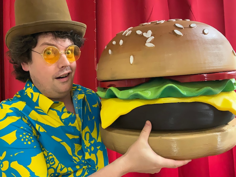 Puede incluir: Una persona con un sombrero marr&oacute;n, gafas de sol amarillas y una camisa con estampado azul y amarillo est&aacute; sosteniendo una gran hamburguesa marr&oacute;n con lechuga, queso y un tomate rojo.