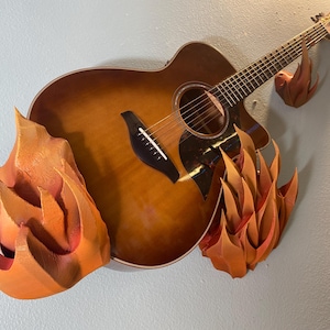 Puede incluir: Una guitarra acústica marrón con una protección negra está montada en una pared con tres ganchos de pared en forma de llama naranja.