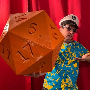 Pu&ograve; includere: Grande dado D20 arancione con numeri bianchi. Il dado &egrave; tenuto da una persona che indossa un cappello da capitano, occhiali da sole e una camicia floreale blu e gialla. Lo sfondo &egrave; una tenda rossa.