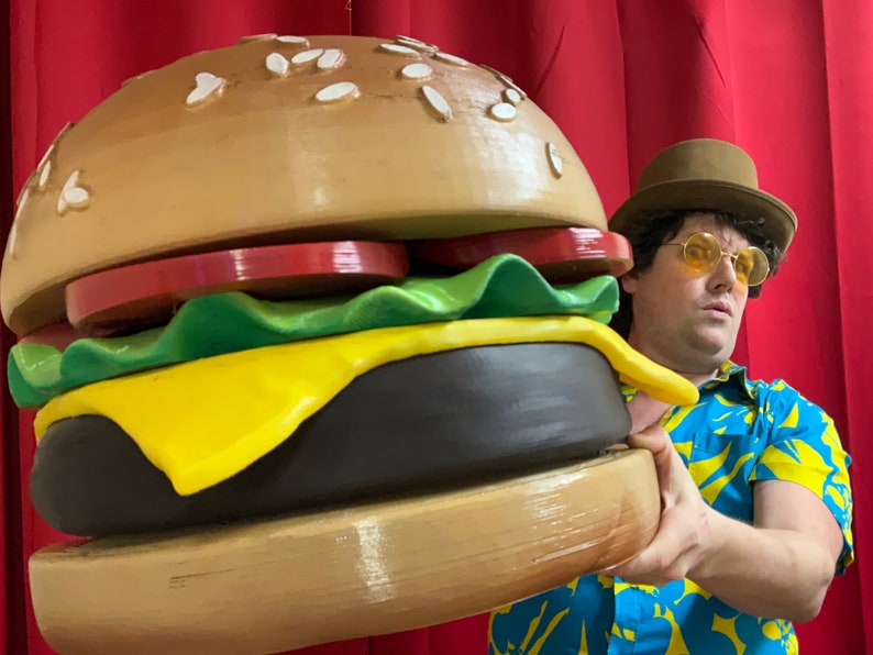 Puede incluir: Una persona con una camisa hawaiana azul y amarilla y un fedora marr&oacute;n sostiene una gran hamburguesa de pl&aacute;stico de gran tama&ntilde;o. La hamburguesa est&aacute; hecha de pl&aacute;stico marr&oacute;n, amarillo, verde y rojo.