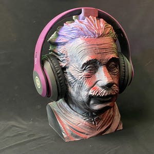 May include: A bust of Albert Einstein with a pair of purple and black headphones resting on his head. The bust is painted in shades of red, gray, and blue. The headphones have the word "LOGO" printed on the side.