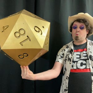 Pu&ograve; includere: Un grande dado a venti facce color oro, con i numeri 4, 6, 8, 9 e 18 visibili. Il dado &egrave; tenuto da una persona che indossa una maglietta nera con testo bianco e una striscia rossa, una camicia bianca floreale, un cappello da cowboy e occhiali da sole.