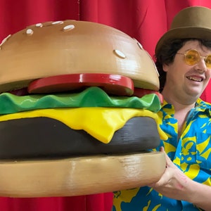 Puede incluir: Una persona sosteniendo una gran hamburguesa marr&oacute;n y amarilla con lechuga, tomate y queso. La hamburguesa es mucho m&aacute;s grande que la cabeza de la persona.