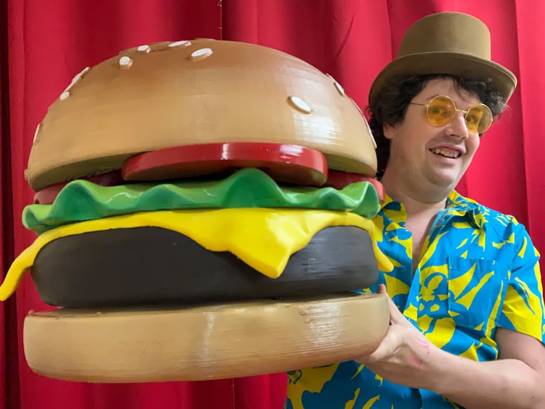 Puede incluir: Una persona sosteniendo una gran hamburguesa marr&oacute;n y amarilla con lechuga, tomate y queso. La hamburguesa es mucho m&aacute;s grande que la cabeza de la persona.
