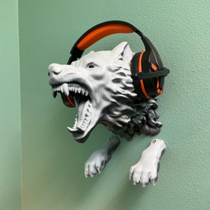 Könnte beinhalten: Ein 3D-gedruckter Wolfskopf mit offenem Maul und scharfen Zähnen, der einen schwarzen und orangefarbenen Kopfhörer hält. Der Wolfskopf ist an einer Wand mit zwei Pfotenabdrücken auf beiden Seiten befestigt.