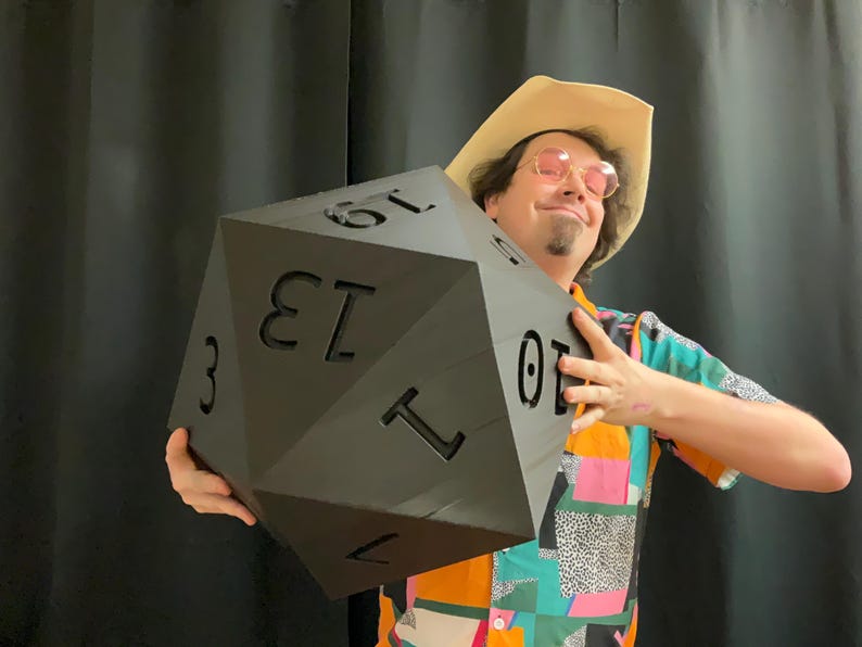 Pu&ograve; includere: Un grande dado nero a venti facce con numeri bianchi. Il dado &egrave; tenuto da una persona che indossa un cappello da cowboy e una camicia colorata. I numeri 1, 3, 7, 10, 13 e 19 sono visibili sul dado.