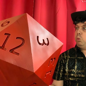 Puede incluir: Un dado rojo grande de 12 caras con los n&uacute;meros 1, 2, 3, 6, 12 y 3 impresos en las caras visibles. Una persona con un sombrero negro y una camisa con estampado negro y dorado est&aacute; sosteniendo el dado.