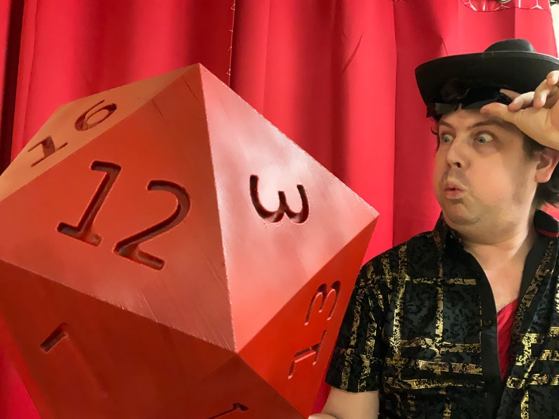 Puede incluir: Un dado rojo grande de 12 caras con los n&uacute;meros 1, 2, 3, 6, 12 y 3 impresos en las caras visibles. Una persona con un sombrero negro y una camisa con estampado negro y dorado est&aacute; sosteniendo el dado.