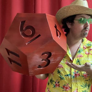 May include: A large, red, wooden D20 die with carved numbers 3, 6, 11, and 16. The die is held by a person wearing a straw hat, green sunglasses, and a yellow Hawaiian shirt with a flamingo pattern. The background is a red curtain.