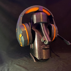May include: A black and orange gaming headset rests on a 3D-printed Moai head stand. The headset features orange accents and a microphone. The stand is a dark, textured color, resembling stone. The overall theme is gaming accessories.
