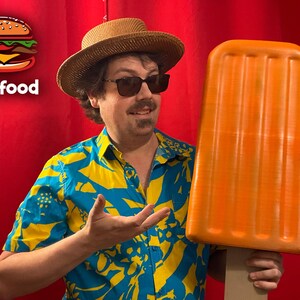 Könnte beinhalten: Eine Person hält eine große orangefarbene Eis am Stiel-Requisite. Die Person trägt einen Strohhut, eine Sonnenbrille und ein blau-gelb gemustertes Hemd. Das Bild enthält den Text "big food" mit einer Burger-Grafik.