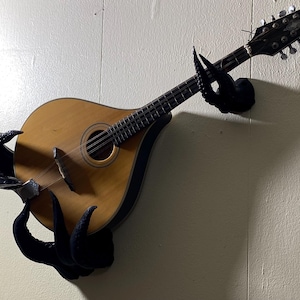 Peut inclure: Un mandoline fixée au mur, maintenue par un support noir stylisé en forme de tentacule. La mandoline a un corps en bois brun clair et une touche sombre. Le mur est peint en beige clair.
