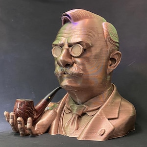 May include: A bronze-colored bust of a man with a mustache, round glasses, and a pipe. The sculpture is detailed, showing the man's suit, tie, and hair. The pipe is held in the man's hand. The bust is a decorative item.