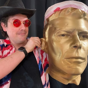 Peut inclure: Une personne portant un chapeau de cowboy et des lunettes de soleil rouges tient un buste en or imprimé en 3D d'une personne. Le buste porte un bandeau en plastique blanc.