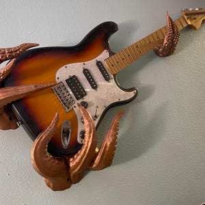 Może przedstawiać: Gitara elektryczna z wykończeniem sunburst i białym pickguardem jest ozdobiona brązowymi mackami ośmiornicy. Macki owijają się wokół korpusu i główki gitary, tworząc unikalne dzieło sztuki ściennej.