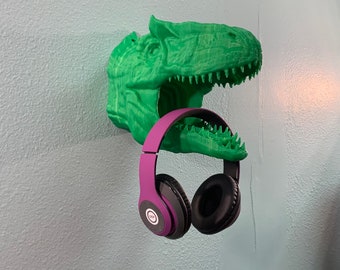 May include: A green 3D printed dinosaur head mounted on a wall with a pair of purple headphones hanging from its mouth.