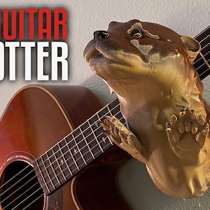 May include: A golden brown otter figurine is mounted on a wooden acoustic guitar. The guitar has a natural wood finish and a black fretboard. The otter is looking up and has a playful expression.