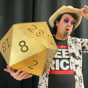 Pu&ograve; includere: Un grande dado a venti facce color oro con numeri neri. Il dado &egrave; tenuto da una persona che indossa un cappello di paglia, occhiali da sole e una maglietta nera con la scritta "SEE RICH" in lettere bianche su una striscia rossa.