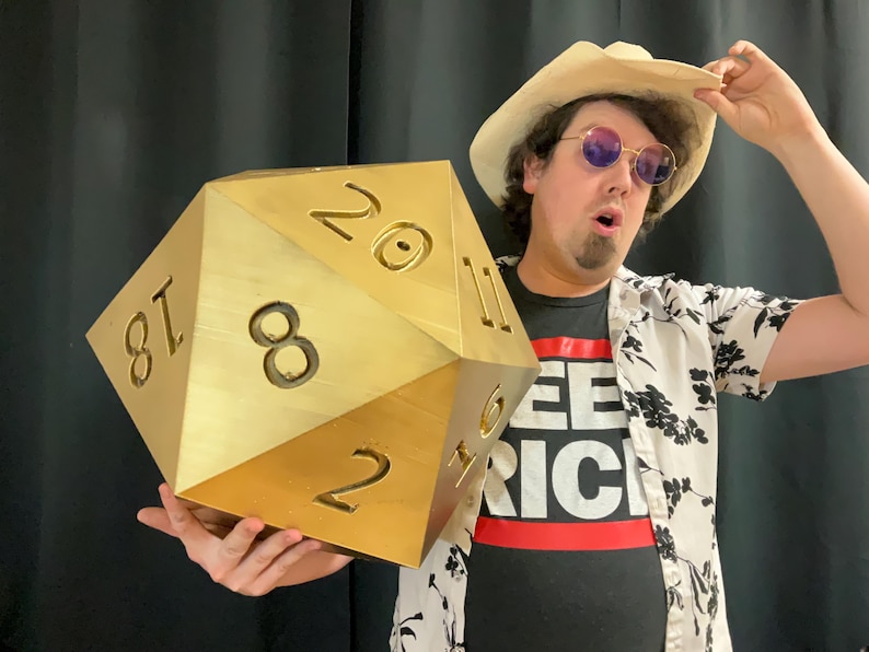 Pu&ograve; includere: Un grande dado a venti facce color oro con numeri neri. Il dado &egrave; tenuto da una persona che indossa un cappello di paglia, occhiali da sole e una maglietta nera con la scritta "SEE RICH" in lettere bianche su una striscia rossa.