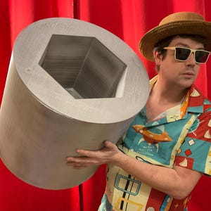 May include: A large, cylindrical, silver-colored object with a hexagonal interior. The object is being held. The background is a solid red curtain. The person holding the object is wearing a patterned shirt, sunglasses, and a straw hat.