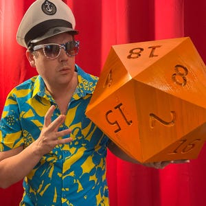 Pu&ograve; includere: Una persona tiene in mano un grande dado arancione a venti facce con numeri neri. La persona indossa un cappello da capitano, occhiali da sole e una camicia a fiori blu e gialla. Il dado &egrave; un accessorio per un gioco da tavolo.