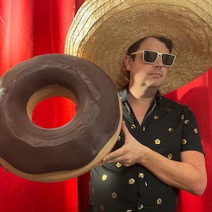 Peut inclure: Un grand beignet glacé au chocolat est tenu devant une personne portant un sombrero de paille et des lunettes de soleil. Le beignet a une base marron clair et un glaçage marron foncé. La personne porte une chemise noire avec des ornements dorés.