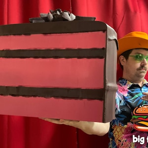 Puede incluir: Una persona con un sombrero de vaquero amarillo y una camisa colorida sostiene un gran pastel de chocolate de tres pisos con glaseado rosa. El pastel está decorado con virutas de chocolate. El texto "big food" está impreso en la parte inferior de la imagen.