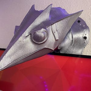 Puede incluir: Un casco plateado impreso en 3D con un diseño angular estilizado. El casco presenta elementos puntiagudos y un detalle central redondeado, lo que sugiere un tema futurista o de fantasía. El casco está apoyado sobre una superficie roja.