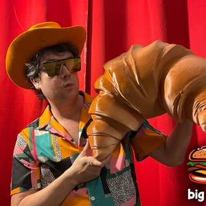 Peut inclure: Une personne portant un chapeau de cow-boy jaune et des lunettes de soleil tient une grande réplique de croissant brun. La personne porte une chemise à motifs colorés. Un logo "big food" avec un burger est dans le coin inférieur droit.