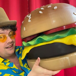 Puede incluir: Una persona con un sombrero marr&oacute;n y gafas de sol sostiene una gran hamburguesa marr&oacute;n y amarilla. La hamburguesa tiene lechuga, tomate y queso.