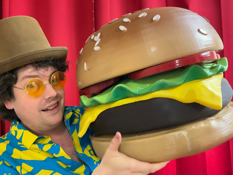 Puede incluir: Una persona con un sombrero marr&oacute;n y gafas de sol sostiene una gran hamburguesa marr&oacute;n y amarilla. La hamburguesa tiene lechuga, tomate y queso.