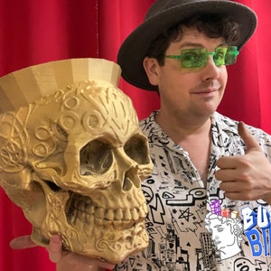 May include: A person wearing a black hat and green glasses holds a gold skull planter. The planter has intricate details and a textured surface. The person is giving a thumbs up and the text "BUST BINS" is visible in the lower right corner of the image.