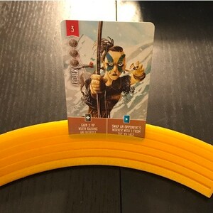 May include: A card game card with an illustration of a woman with blue face paint holding a bow and arrow. The card is titled "Archer" and has a value of 3. The card text reads "Gain 2 VP when raiding an outpost" and "Swap an opponent's worker with 1 from the village."