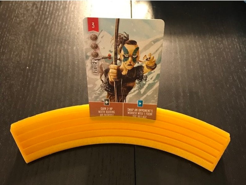 May include: A card game card with an illustration of a woman with blue face paint holding a bow and arrow. The card is titled "Archer" and has a value of 3. The card text reads "Gain 2 VP when raiding an outpost" and "Swap an opponent's worker with 1 from the village."
