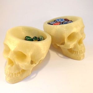 May include: Two off-white ceramic skull-shaped bowls. One bowl is filled with green and blue dice.