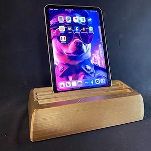 Puede incluir: Un soporte de impresión 3D de color dorado con una ranura para una tableta. Una tableta con una pantalla colorida está en el soporte. La pantalla muestra un perro con gafas de sol y un sombrero de policía.