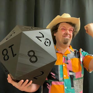 Pu&ograve; includere: Un grande dado nero a venti facce, tenuto da una persona con un cappello da cowboy e una camicia colorata. Il dado ha grandi numeri bianchi. La persona sorride e flette il braccio.