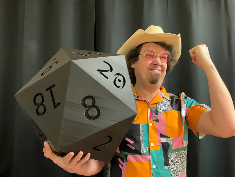 Pu&ograve; includere: Un grande dado nero a venti facce, tenuto da una persona con un cappello da cowboy e una camicia colorata. Il dado ha grandi numeri bianchi. La persona sorride e flette il braccio.